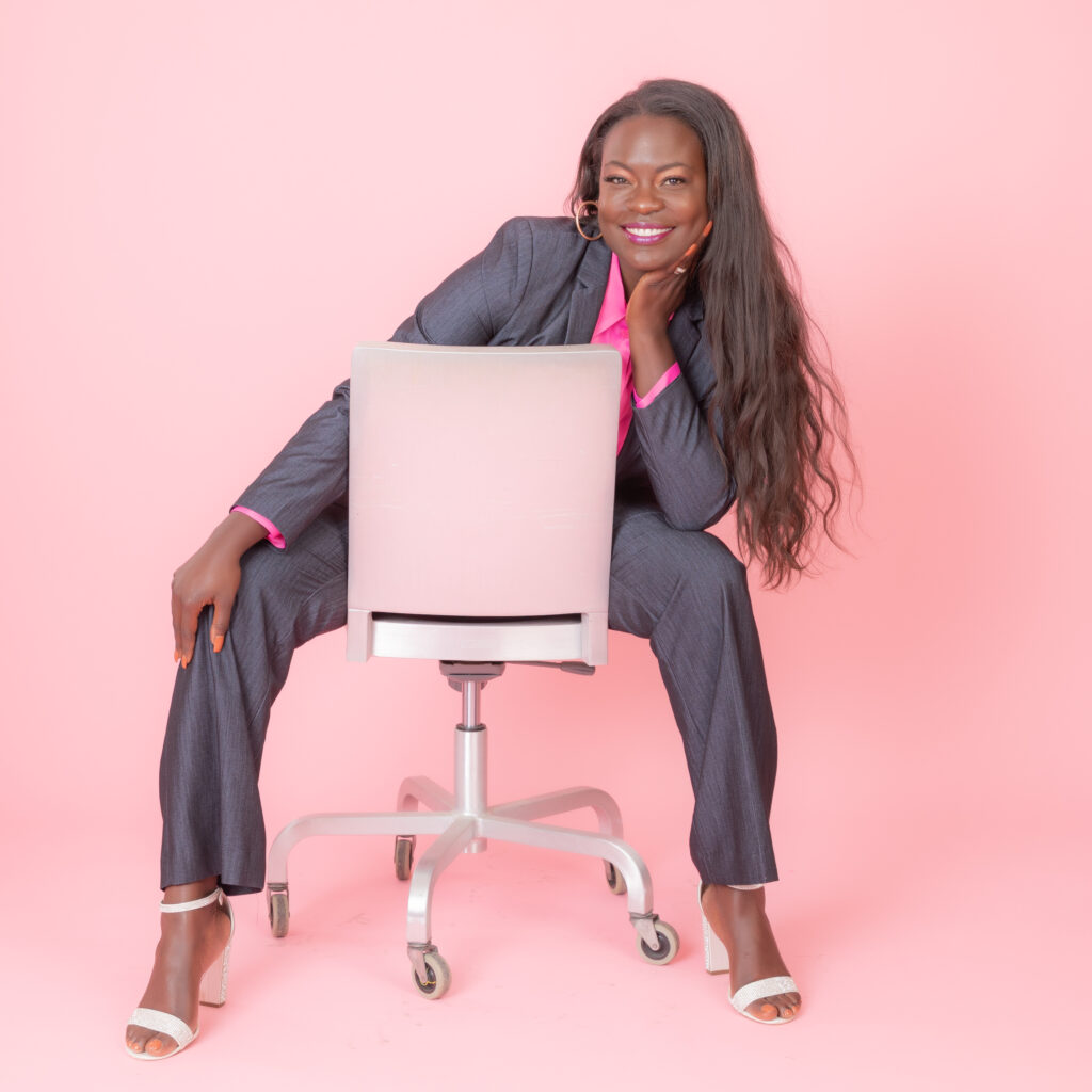 Woman sitting backwards in a chair, resting her face on her hand. She has a pink background behind her.